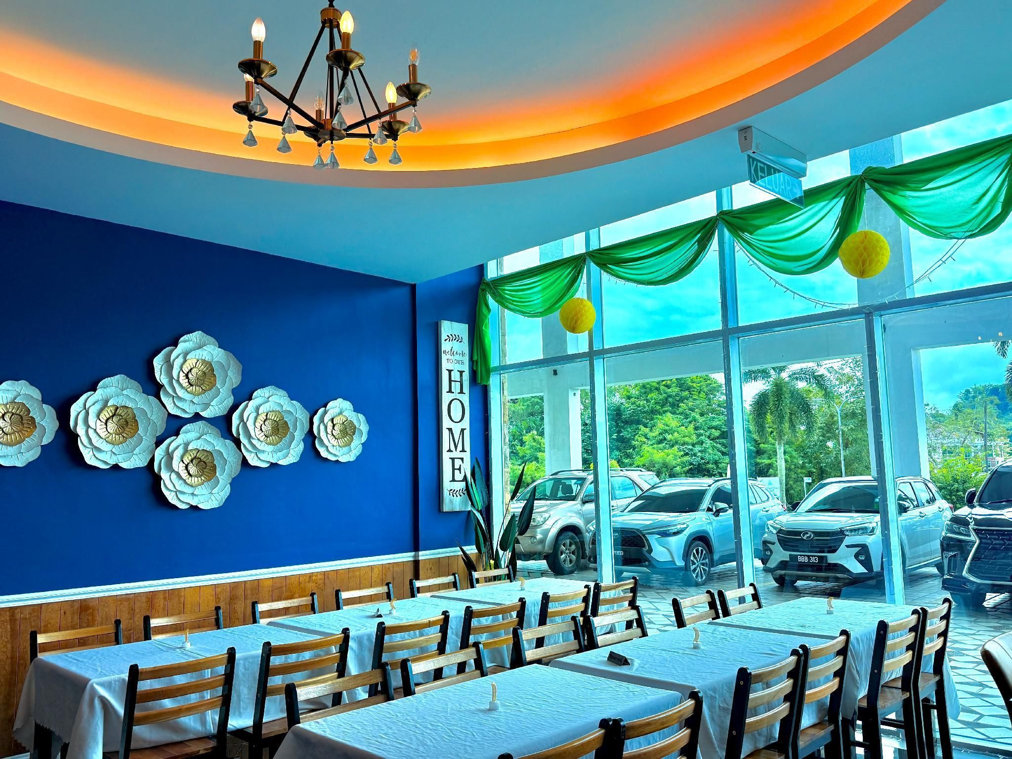 A restaurant with blue walls, white flower decorations, and a 'Welcome to our HOME' sign. Tables are arranged for dining.