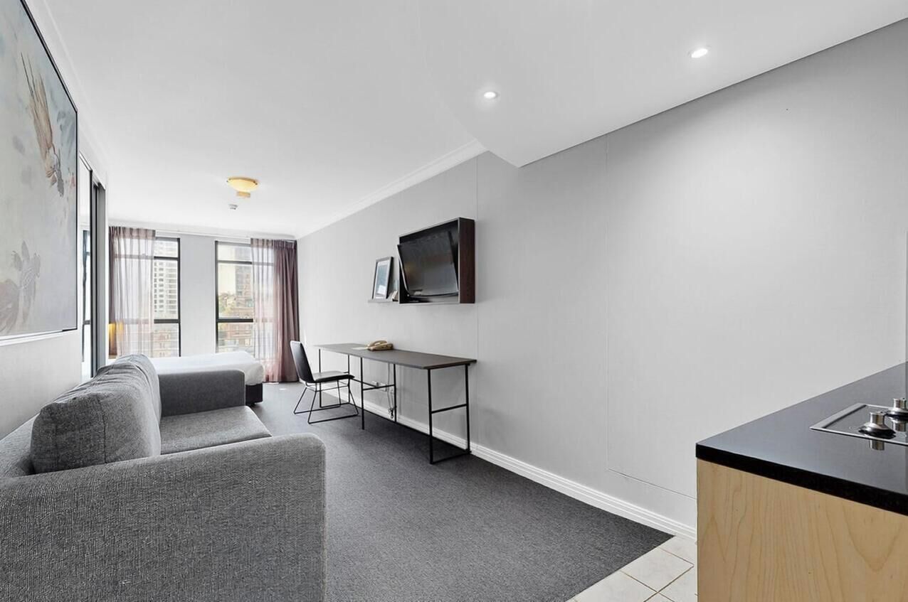 A modern hotel room with a grey sofa, a small desk with a chair, a flat-screen TV mounted on the wall, and a kitchenette area.