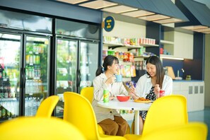A dining area with yellow chairs and tables, a refrigerator with beverages, and shelves stocked with snacks. - Go Hotel Si Racha At Central Si Racha (Si Racha)