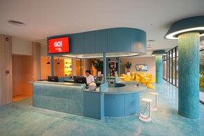Ein Hotel-Rezeptionsbereich mit einer gebogenen, blauen Theke, einem digitalen Bildschirm, der den Hotelnamen anzeigt, und einer Sitzecke mit gelben Stühlen.