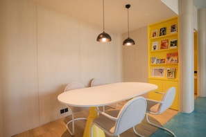 A modern dining area with a white table and chairs, black pendant lights, and a yellow bookshelf with books and magazines. - Go Hotel Si Racha At Central Si Racha (Si Racha)