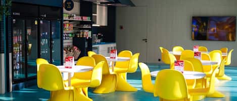 A modern dining area with yellow chairs and white tables, a refrigerated display case, and a wall-mounted television.