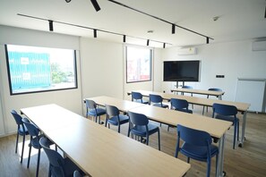 A classroom with long tables and blue chairs, a large screen, and windows with views of buildings. - Go Hotel Si Racha At Central Si Racha (Si Racha)