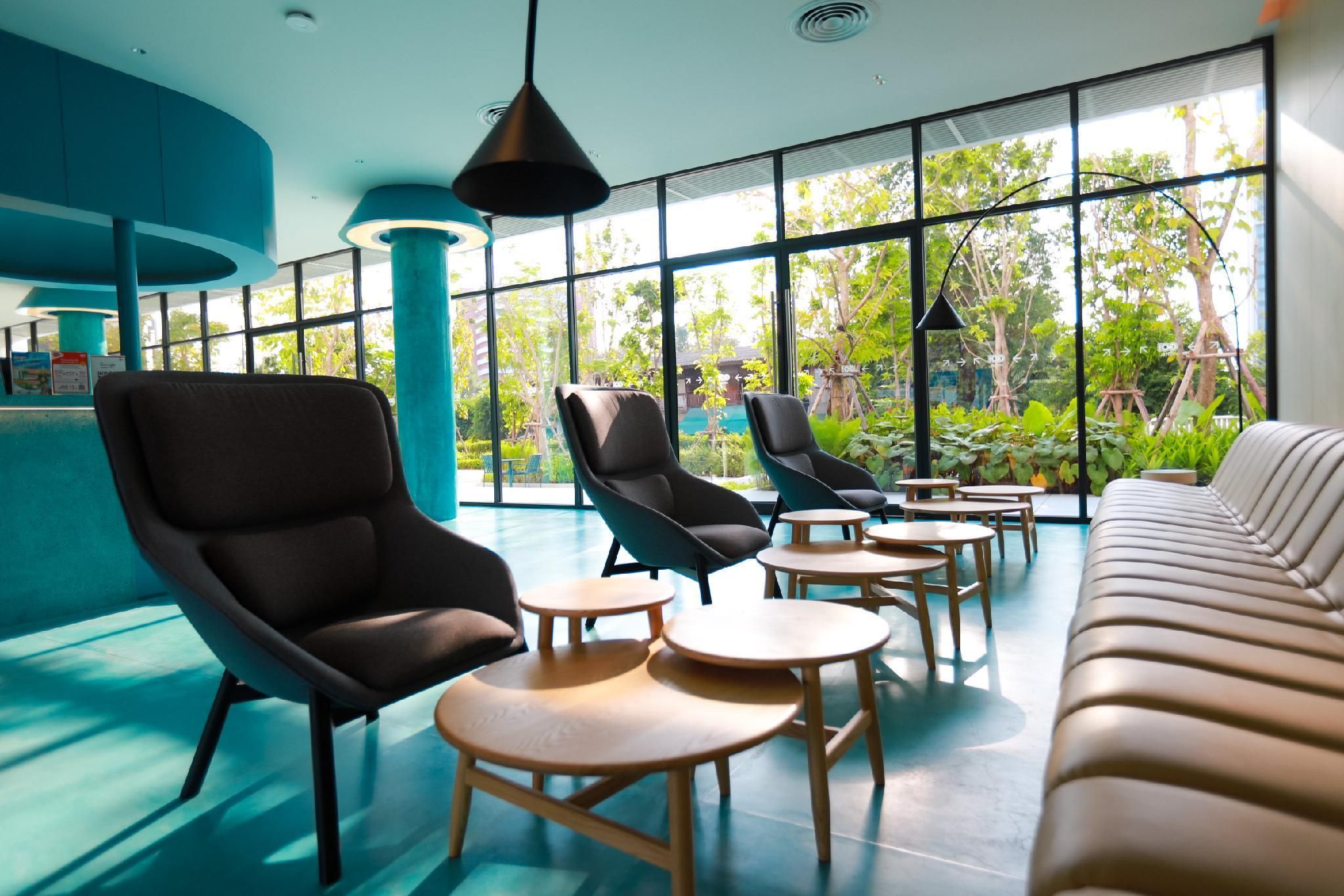 A modern lounge area with black leather chairs, round wooden tables, and a beige sofa. Large windows offer views of greenery outside.