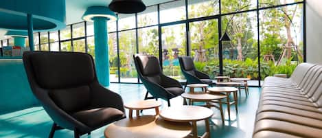 A modern lounge area with black leather chairs, round wooden tables, and a beige sofa. Large windows offer views of greenery outside.