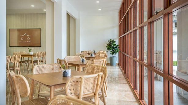 A hotel dining area with wooden tables and chairs, a large potted plant, and a wall-mounted sign. - Kio Hotel Korat (Nakhon Ratchasima)