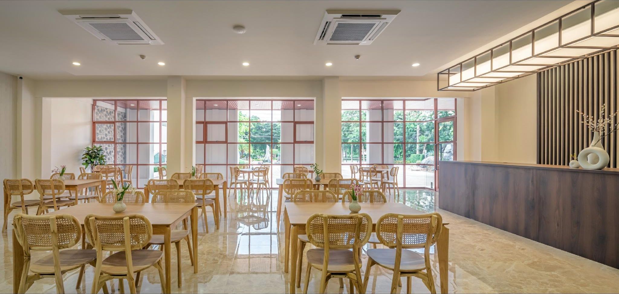 Uma ampla sala de jantar com várias mesas e cadeiras, grandes janelas com vista para áreas verdes, e design moderno com detalhes em madeira e um painel decorativo na parede.