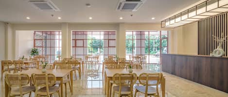 Uma ampla sala de jantar com várias mesas e cadeiras, grandes janelas com vista para áreas verdes, e design moderno com detalhes em madeira e um painel decorativo na parede.