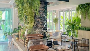 A modern cafe with a central counter, surrounded by seating, and adorned with greenery.