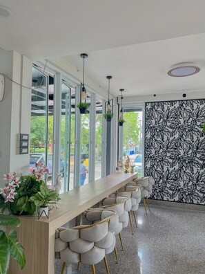 Un'area bar moderna con un lungo bancone in legno, sgabelli imbottiti, lampade a sospensione e un'ampia vetrata che si affaccia sulla strada.