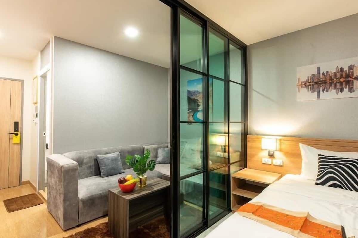 A hotel room with a sofa, a small table with fruit, a bed, a nightstand with a lamp, and a cityscape painting on the wall.
