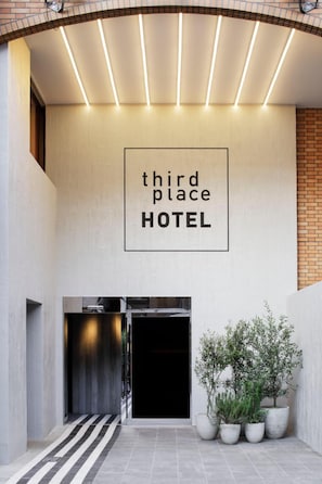 L’entrée d’un hôtel, avec une enseigne indiquant « THIRD PLACE HOTEL » et une composition florale dans un pot.
