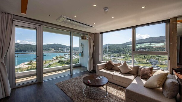 A spacious living room with a large sofa, a coffee table, and a view of a lake and mountains. - Ramada Suites Geoje (Pyeongtaek)