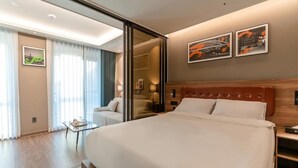 A modern hotel room with a large bed, a glass coffee table, a sofa, and a view of the city through the window.