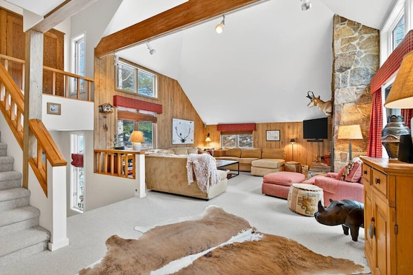 A spacious loft with a high ceiling, exposed wooden beams, a stone fireplace, and a cozy seating area with a sofa and ottoman.