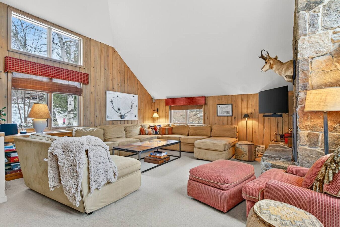 A cozy living room with a stone fireplace, wooden walls, and a large window overlooking a snowy landscape.
