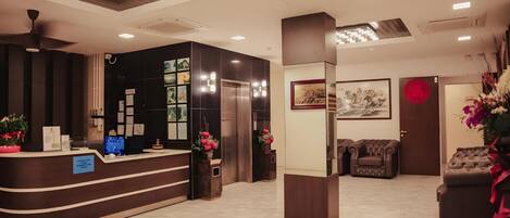 A reception area with a curved desk, a digital screen, and a reception counter. There is a wall-mounted elevator and a door with a red circular sign.