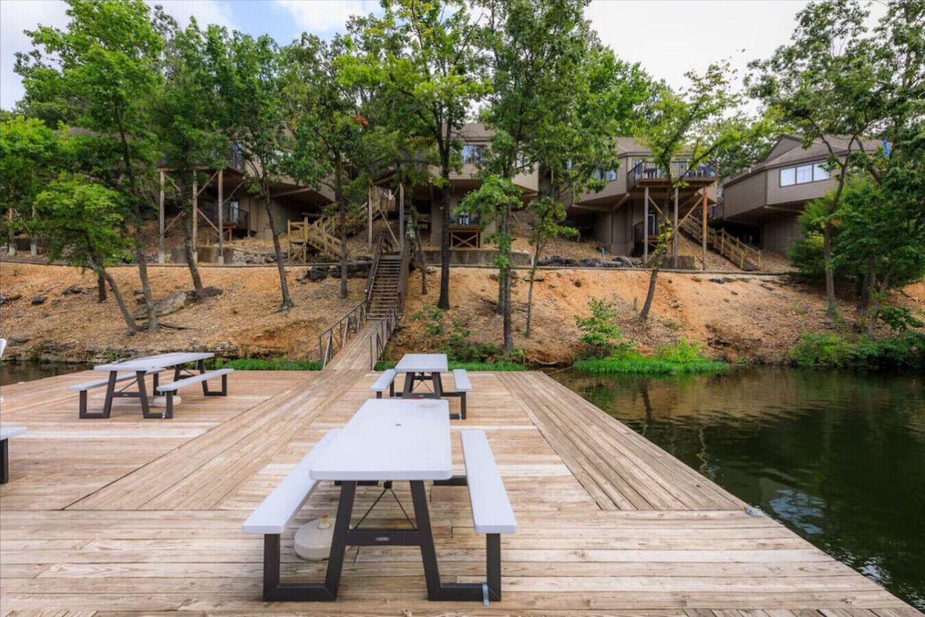 A wooden deck with picnic tables overlooking a body of water, surrounded by trees and buildings.