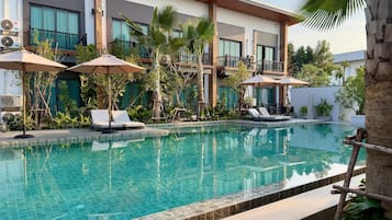 Piscina com espreguiçadeiras e guarda-sóis, cercada por vegetação e um edifício moderno com sacadas.