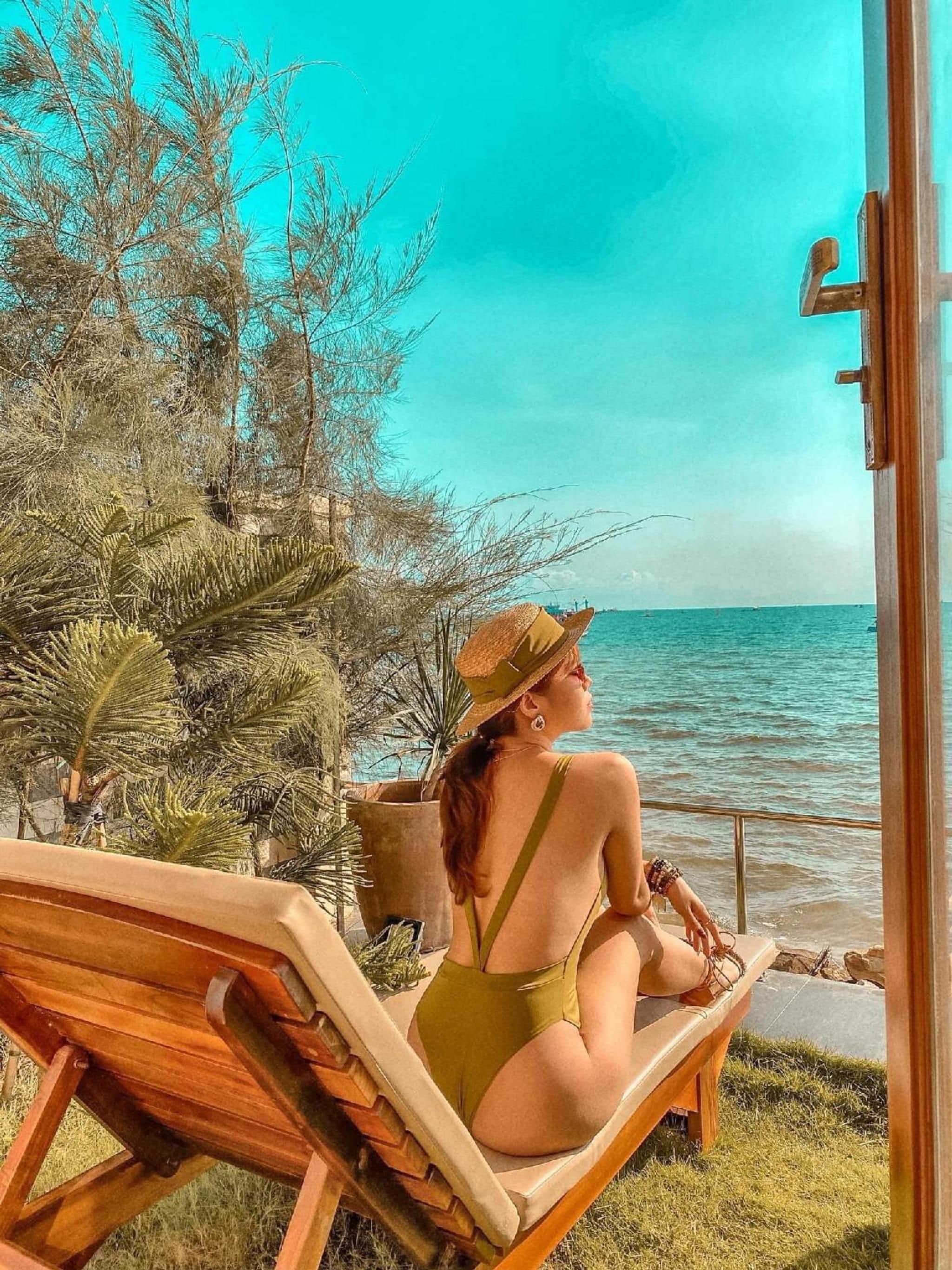 Una mujer sentada en una silla de madera junto al mar, vestida con un traje de baño verde y un sombrero de paja.