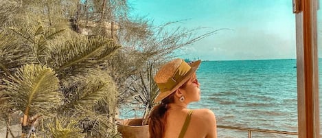 Una mujer sentada en una silla de madera junto al mar, vestida con un traje de baño verde y un sombrero de paja.