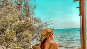 Una mujer sentada en una silla de madera junto al mar, vestida con un traje de baño verde y un sombrero de paja.