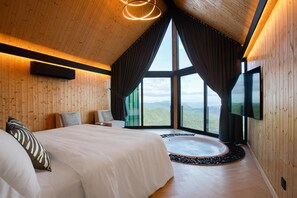 A bedroom with a large bed, a TV, a window with curtains, and a view of the mountains.