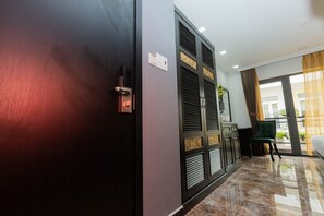 A hotel room with a dark wooden door, a minibar, a desk with a chair, and a view of the outdoors through a window with curtains. - Collection O Havana Airport Hotel (Ho Chi Minh City)