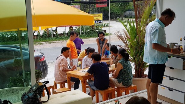 A group of people seated at tables under a yellow umbrella, with a man standing by a counter. - Wangmaewmeo (Rayong)
