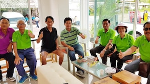 A group of people sitting in a modern indoor setting with glass walls, wooden furniture, and a small table with a cup and snacks. - Wangmaewmeo (Rayong)