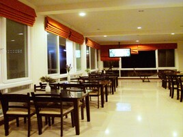 Uma ampla sala de jantar com várias mesas e cadeiras, janelas grandes com cortinas e uma tela de televisão.