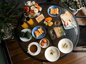 A round glass table with a variety of food items, including rice bowls, a cup of coffee, and a floral arrangement. - The Sky Roof Homestay Kiriwong (Lan Saka)