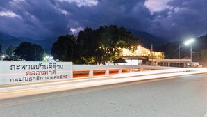Une route avec un panneau en thaï, un bâtiment illuminé et des arbres en arrière-plan.