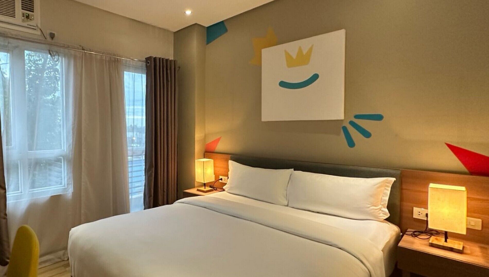 A hotel room with a large bed, two bedside tables with lamps, a window with curtains, and a wall with a simple decorative piece.