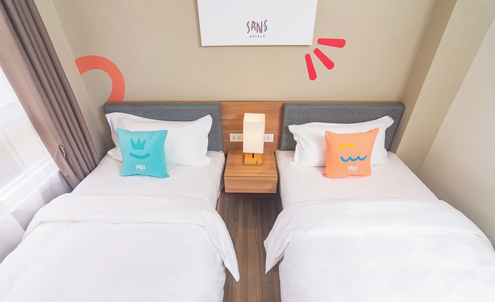 Ein Hotelzimmer mit zwei Betten, jeweils mit weißer Bettdecke und passenden Kissen, einem Nachttisch aus Holz mit Lampe sowie einem an der Wand befestigten Schild über den Betten.