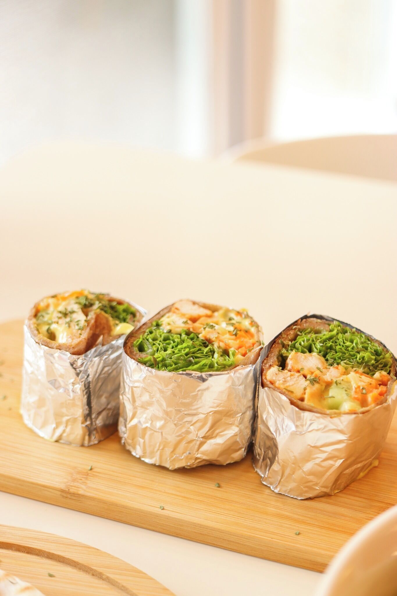 Trois rouleaux de sushis enveloppés dans du papier d’aluminium, posés sur une planche à découper en bois.