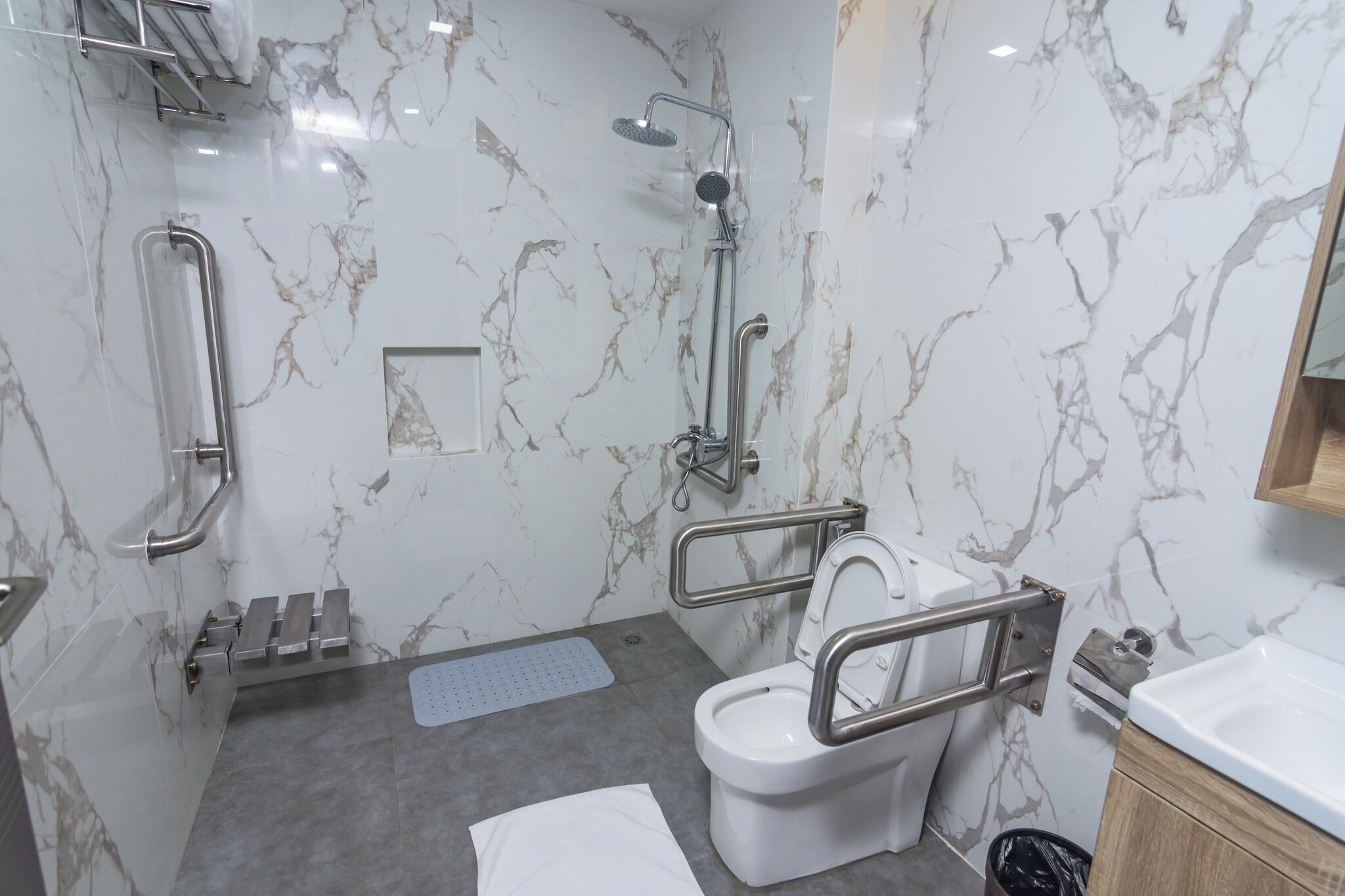 Un baño con pared de mármol, un inodoro blanco con barra de apoyo, una ducha con cabezal de ducha de mano y un lavabo con espejo.