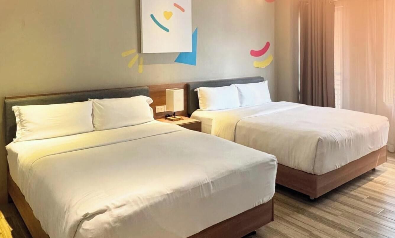 Una habitación de hotel con dos camas, una mesita de noche con lámpara y una pared decorada con arte abstracto.
