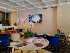 Un hall d’hôtel avec une table ronde, des chaises bleues, un pot d’orchidée, des magazines et un téléviseur fixé au mur.