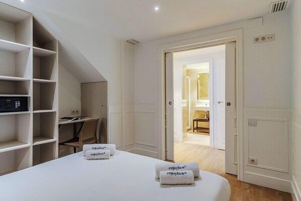 Quarto de hotel com cama, escrivaninha e estante. Há toalhas na cama e uma porta que dá para o banheiro.