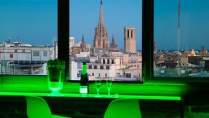 Vista desde una ventana con una zona de bar iluminada en verde, que muestra una botella y vasos, con vistas a un paisaje urbano donde destaca una catedral.