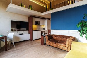 Un salon moderne avec un canapé en cuir marron, un fauteuil jaune, un téléviseur à écran plat, un coin cuisine équipé d’un four à micro-ondes et un escalier menant à un étage supérieur.