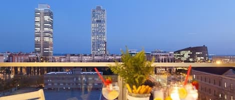 A balcony with a table set for two, overlooking a cityscape with modern buildings and a waterfront.