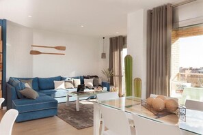 Een moderne woonkamer met een blauwe bank, een glazen salontafel en uitzicht op de stad door grote ramen.