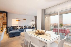 A modern living room with a glass dining table, a blue sofa, and a city view through large windows. - Enjoybcn Marina Apartment (Barcelona)