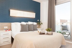 Un dormitorio moderno con pared de ladrillo azul, una cama grande con ropa de cama blanca y gris, una mesita de noche con libros, una bandeja con una taza y flores, y vistas de la ciudad a través de un ventanal grande.