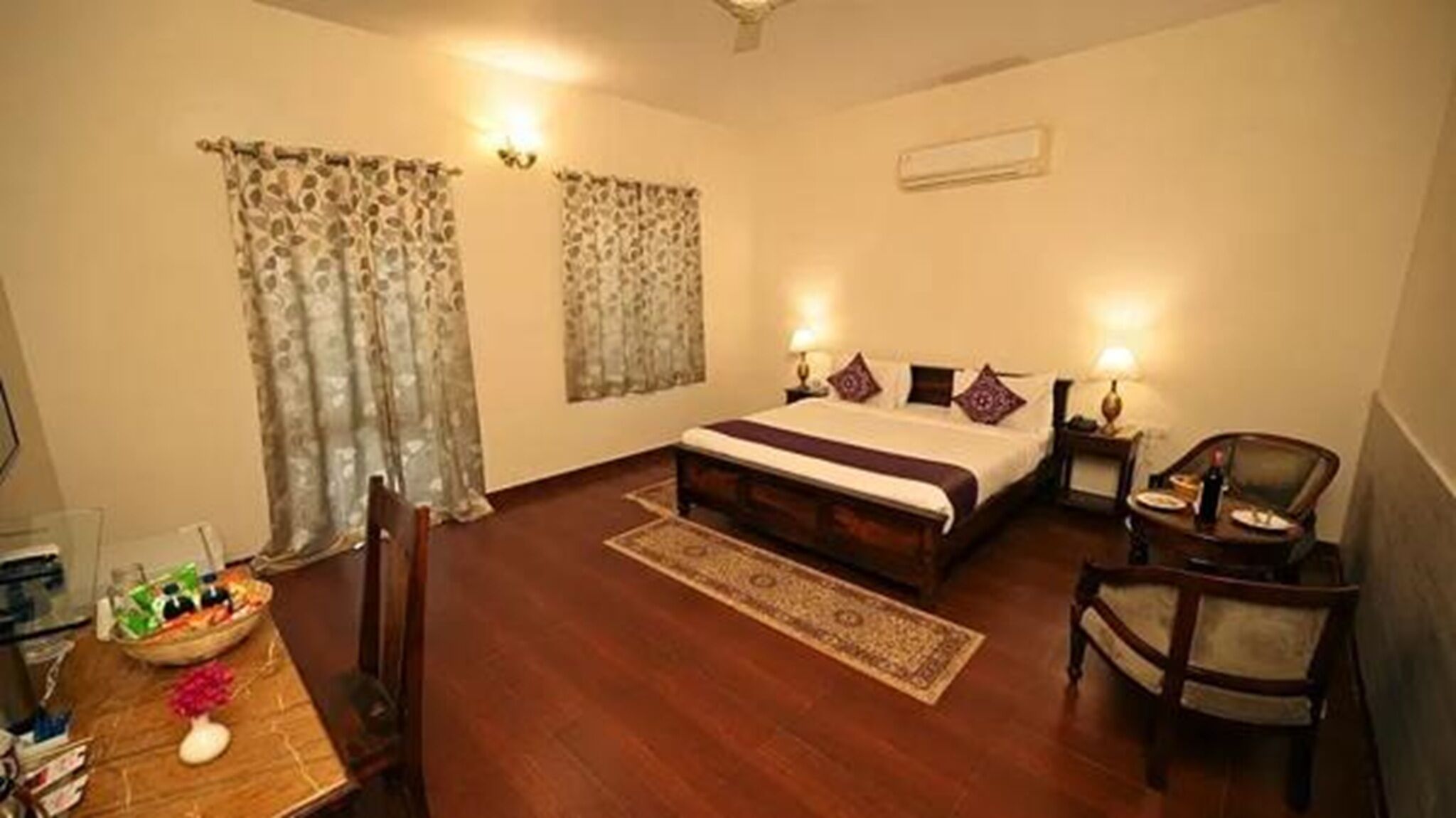 A hotel room with a wooden bed, two bedside tables with lamps, a chair, a small table with a vase, and a rug.