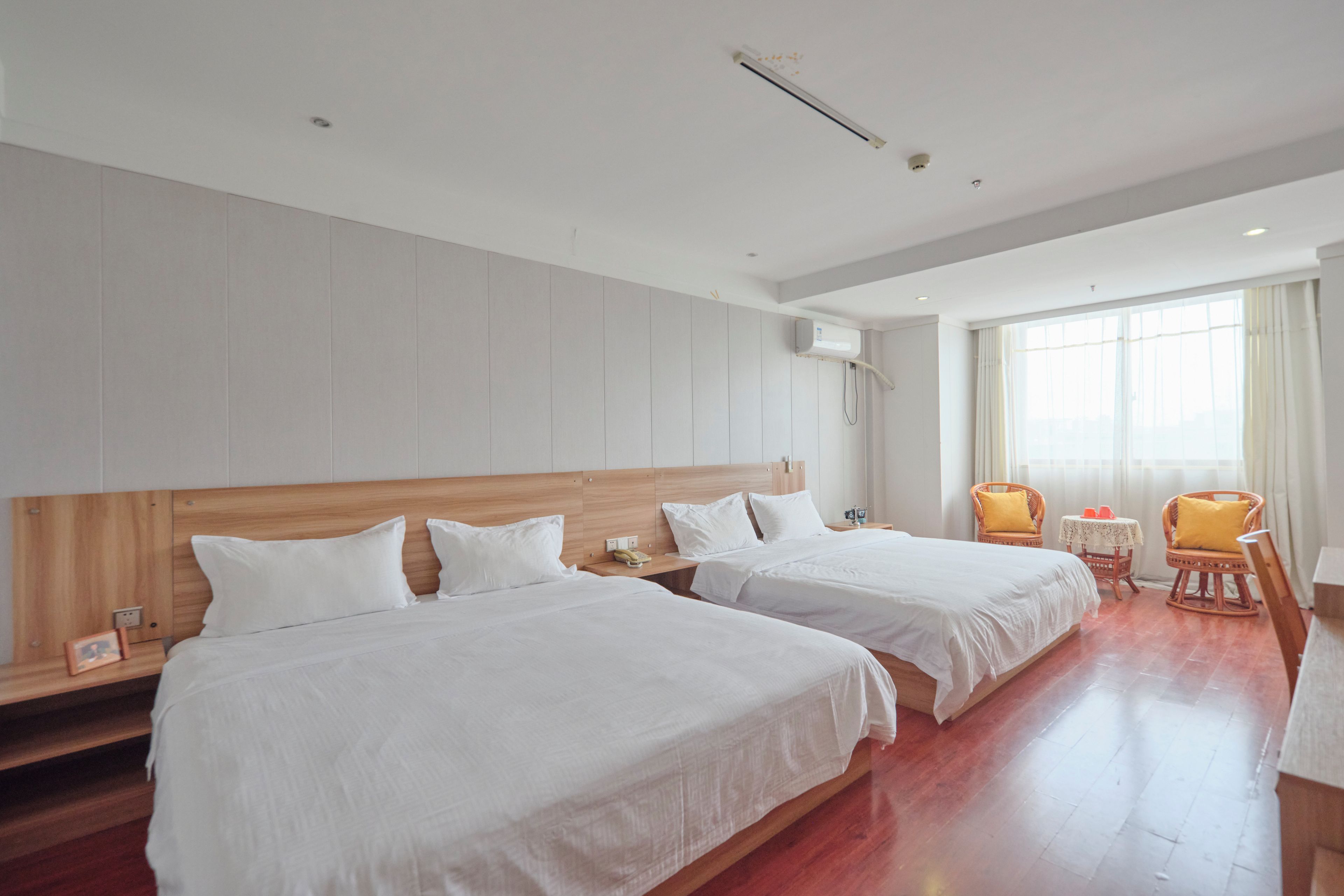 Une chambre d’hôtel avec deux lits, des têtes de lit en bois, une petite table, deux chaises et une grande fenêtre avec des rideaux.