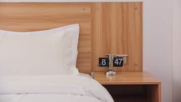A neatly made bed with white bedding and a wooden headboard. A bedside table with a clock displaying the time 8:47 is visible.