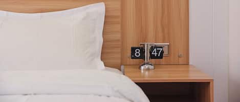 A neatly made bed with white bedding and a wooden headboard. A bedside table with a clock displaying the time 8:47 is visible.